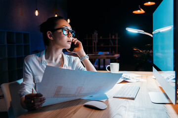 Young businesswoman in eyeglasses holding blueprint and talking on smartphone