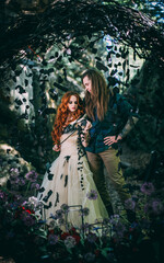 Beautiful young couple together near dark circle arch with flowers. Man with tattoo and long hair with beard. Woman with red hair. Fairytale concept.