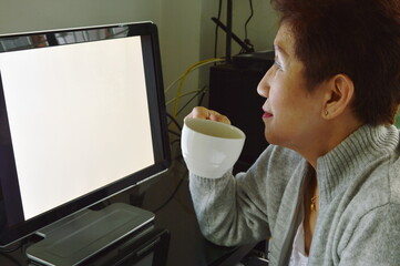 Asian elder woman drinking coffee while working with personal computer