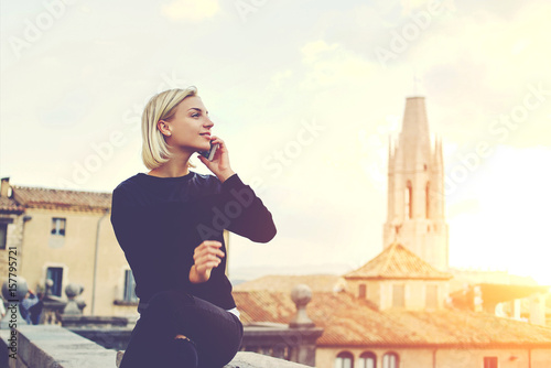Fotografía Young beautiful woman with good mood is talking on mobile phone, while is sitting on a building roof