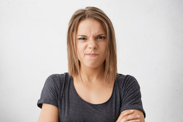 Wall Mural - Angry young female with dark eyes frowning her nose and lips wearing casual T-shirt standing crossed hands isolated over white background showing her dissatisfaction. People and emotions concept
