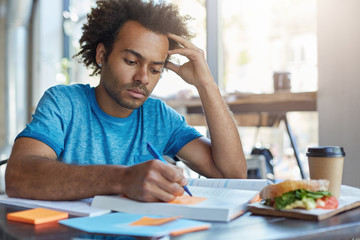 Wall Mural - Handsome serious black European male student busy learning lessons during lunch at cafe, sitting at table with food and textbooks, making notes, writing down new words preparing for Spanish class