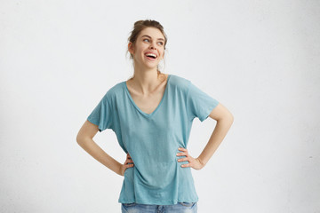 Wall Mural - Isolated portrait of attractive cheerful young European woman wearing oversize blue top smiling broadly, feeling happy and carefree while having fun indoors, looking away with pleasure and joy