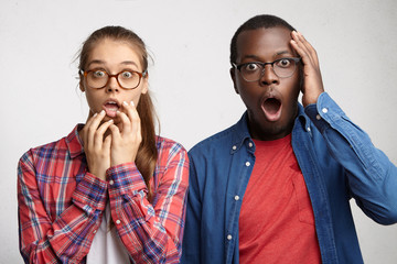 Frightened two people with different races staring at camera with great surprise. Cute female with eyeglasses holding hands on mouth looking with amazement, dark-skinned male holding hand on head