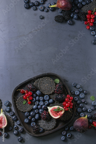 Fotografija Variety of fresh berries blueberry, dewberry, red currant and figs with black charcoal crackers on wooden plate over dark metal background
