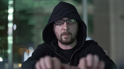 Wall Mural - hacker in hood and eyeglasses typing something on computer in dark room. green lights on screen refl