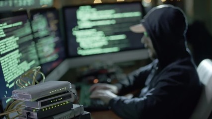 Wall Mural - hooded man typing on keyboard while working at desk with computer workstation in dark room at night