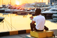 Stylish Man And His Dog Sitting Together On Pier And Enjoying Colorful Sunset. Jack Russell Terrier Puppy On His Lap. Luxury Yachts Docked In Sea Port.