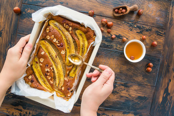 Wall Mural - Homemade fruit banana and hazelnut cake in a baking dish. Woman hands cover the pie with maple syrup. The rustic wooden table, top view.