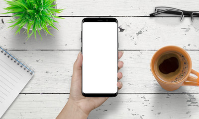 Poster - Smart phone in woman hand with isolated white screen for mockup. Wooden white desk in background with cup of coffee, plant, glasses beside.