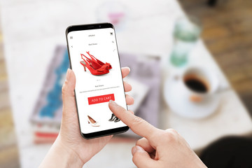 Poster - Add to cart red shoes on online store. Modern smart phone with round edges in woman hand. Table with coffee and magazines in background.
