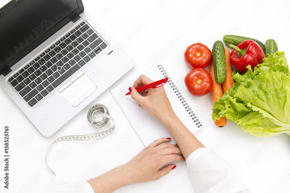 Vegetable diet nutrition or medicaments concept. Doctors hands writing diet plan, ripe vegetable composition, laptop and measuring tape on white background - obrazy, fototapety, plakaty 