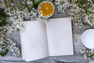 The book with empty pages is surrounded by branches and flowers of white lilac, a mug of tea with lilac petals on a aged wooden vintage background and a white candle in a glass and matches.