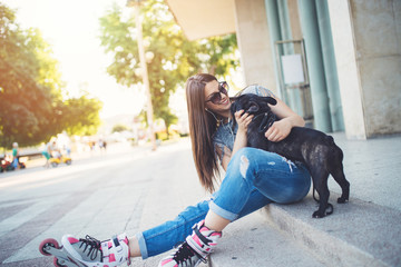 Warm summer colors and haze. Strong back light. Roller skates girl with sitting on city square with her French bulldog and enjoying hot afternoon.