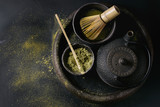 Fototapeta  - Green tea matcha powder and hot drink in black bowls standing with iron teapot, bamboo traditional tools spoon and whisk in terracotta tray over dark metal background. Top view with space