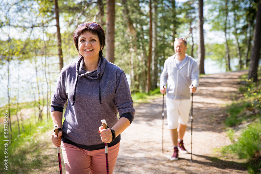 Summer sport in Finland - nordic walking. Man and mature woman hiking in green sunny forest. Active people outdoors. Scenic peaceful Finnish summer landscape. - obrazy, fototapety, plakaty 
