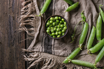 Young organic green pea pods and peas in can tin over old dark wooden planks with sackcloth textile background. Top view with space. Harvest, healthy eating.