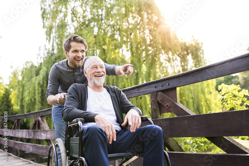 Fotografie, Obraz Hipster son walking with disabled father in wheelchair at park.
