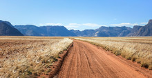 Straight Desert Dirt Road Track Passes Grassland Towards Mountains.