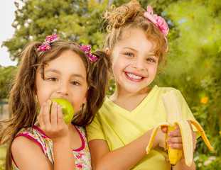 Two beautiful young girls, eating a healthy apple and banana in a garden background