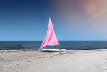 Small White Sailboat With Pink Sails On Empty Beach With Ocean And Vessels On Distant Horizon Under Clear Blue Sky
