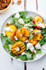 Fresh salad with grilled peach halves, arugula and burrata on a plate on white distressed wooden background. Top view. Summer food concept