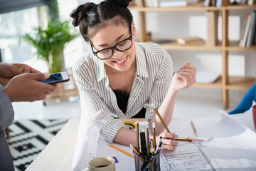 Wall Mural - Smiling young asian businesswoman in eyeglasses working with blueprint in office