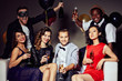 © pressmaster - Group portrait of trendy young people sitting on sofa with champagne flutes in hands and posing for photography with wide smiles, two handsome men standing behind them