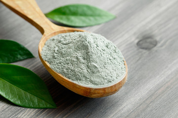 Close-up of a cosmetic dry gray clay in a wooden spoon on a black wooden table. Decorated with green leaves.