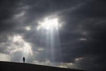 A Man Walking Across Sand Dunes As A Ray Of Light Shines Through The Clouds