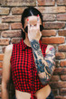 © BONNINSTUDIO/Stocksy - Young alternative woman with tattoos covering her face with a heavy metal sign