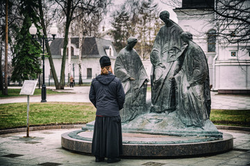 YAROSLAVL, RUSSIA - APRIL 27, 2017: Russian priest in a cassock and a hooded jacket near a sculptural composition Trinity
