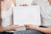 Young Couple Holding A Frame With A Clean Sheet On A White Background. Man And Woman Together. Wedding Invitation , Event Concept. Empty Space For The Text.