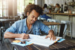 © wayhome.studio  - Stylish hipster guy with African hairstyle wearing denim shirt sitting in cafeteria working with books writing some notes preparing for lessons. Hard-working student being busy with studying