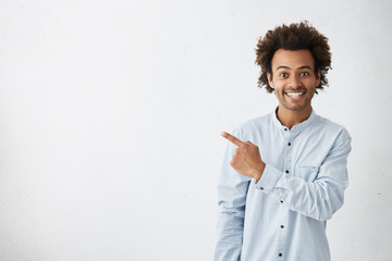 Horizontal portrait of dark-skinned handsome man having broad smile wearing formal white shirt posing against white background pointing with index finger at white copy space for your advertisment