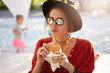 Stylish tourist female wearing summer hat, sunglasses and red shirt having lunch outside eatting sandwich. Young student female tasting fast food outdoors admiring summer vacations. Tourism concept