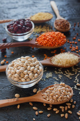 Raw organic cereal grains, seeds and beans (millet, rye,wheat, buckwheat, red and white beans, lentil, rice)  in wooden spoons and bowls on dark stone table, selective focus. Healthy eating concept