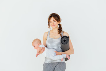 Wall Mural - Sport, motherhood and active lifestyle concept - happy charming sporty mother holding cute little baby boy in one hand and yoga mat in the other while standing on white background.