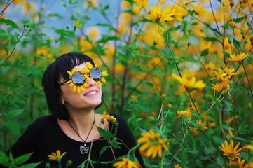 Wonderful portrait of cheerful smiling young brunette woman in sunglasses, out of which Peeps the yellow flower petals are applied to eyes. The girl is happy around Sunny flowers.