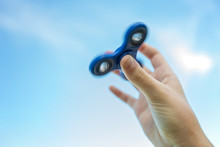Girl's Hand Holding A Spinning Fidget Spinner In Her Hand, Spinning Them On Her Thumb, Against The Blue Sky