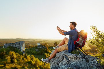 hiking man taking photo with mobile phone sitting on top of the rock
