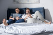 Happy gay couple in eyeglasses lying together in bed and using digital devices