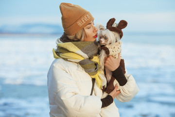 Winter vacation concept. Couple of friends standing on beach near water with ice. Blond girl kissing her Chinese crested dog. Hipster style. Sunny weather. Happy together