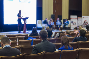 Business Conferences Ideas. Group of People Attending Conference and Listening to the Host Speaker On Stage. Back View of Listeners.