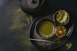 Fototapeta  - Green tea matcha powder and hot drink in black bowls standing with iron teapot, bamboo traditional tools spoon and whisk in vintage tray over dark metal background. Top view with space