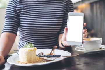 Mockup image of a woman holding and showing white mobile phone with blank screen , cake and coffee cup on wooden table in modern cafe