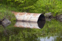 Round Metal Culvert Opening Into Calm Standing Water And Reflecting On The Surface