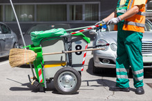 Worker Of Cleaning Company In Green Uniform With Garbage Bin And A Broom. Utility Service Company Men Worker Of Municipality Sweeping City Streets.