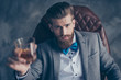 Cheers, ladies and gentlemen! Stylish elegant red bearded aristocrat in suit and bowtie holds glass with brandy, relaxing, sits on leather brown arm chair indoors