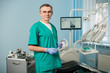 © anatoliy_gleb - Portrait of handsome male dentist at the morden dental office. Doctor wearing green uniform, blue gloves, holding glasses and looking to the camera. Dentistry